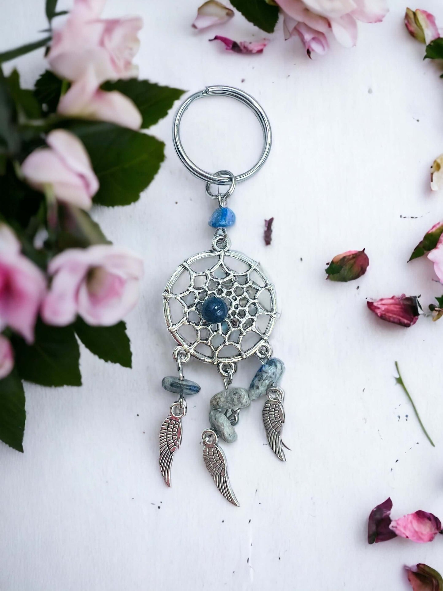 Dream catcher key ring with crystal chips on white surface surrounded with petals.