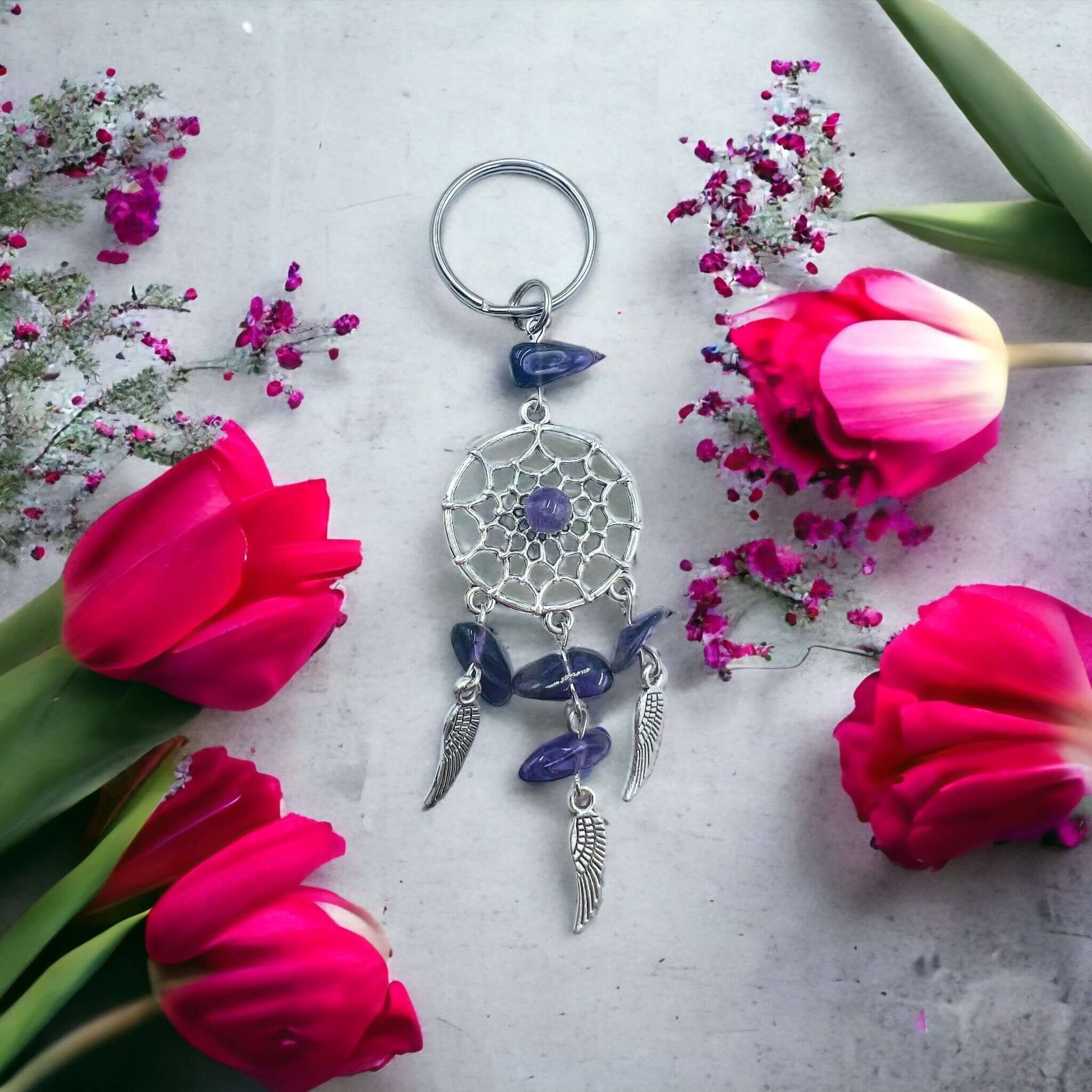 Dream catcher key ring with crystal chips on white surface surrounded with petals. 