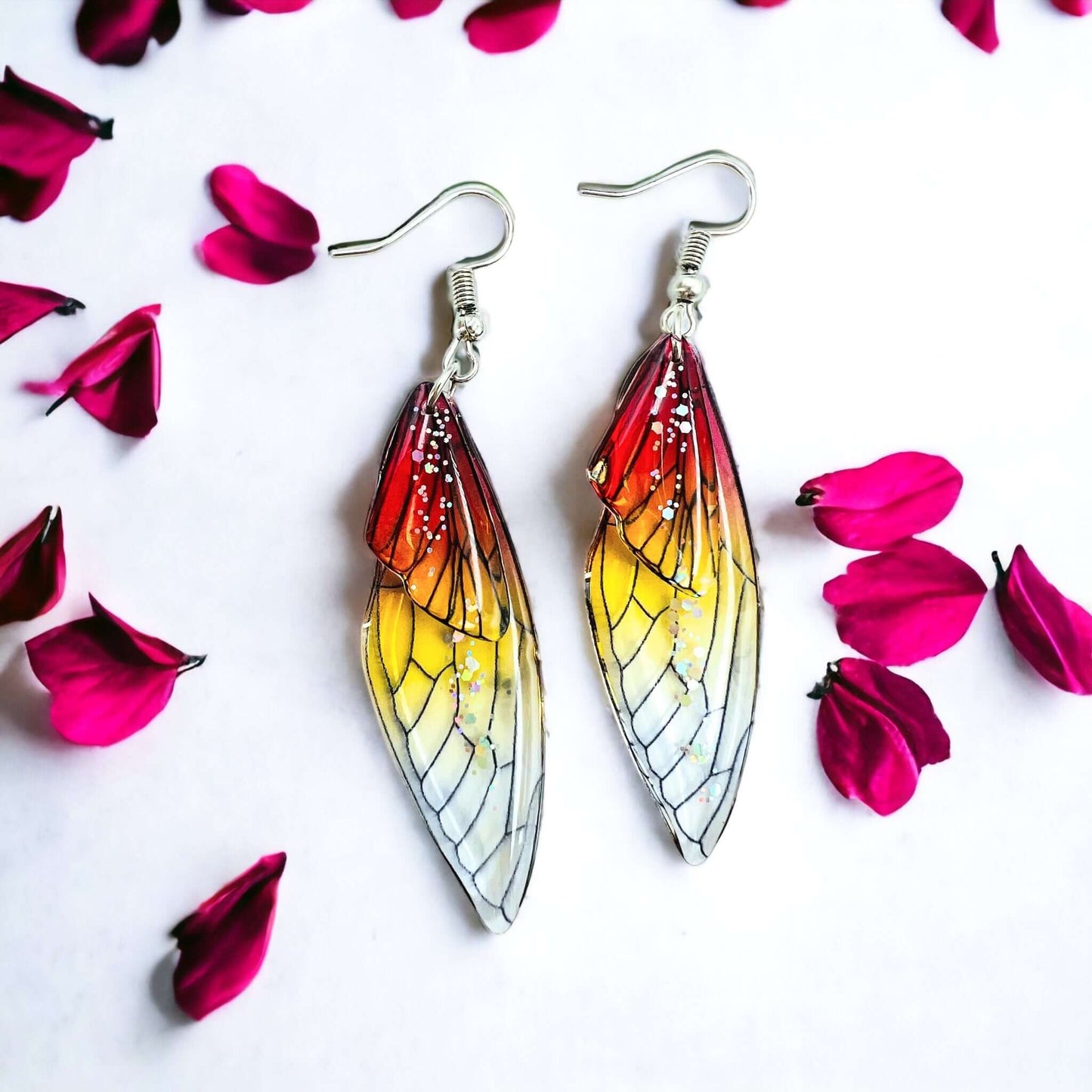 Fairy wing earrings with petals scattered on a white surface.